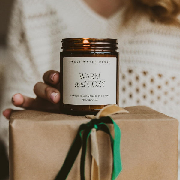 Candle labeled 'Warm and Cozy' held over a wrapped gift with a blurred background