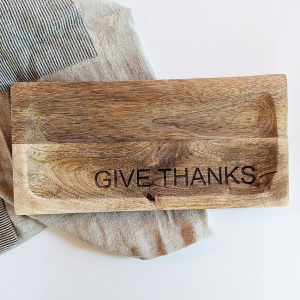 A mango wood serving board with 'Give Thanks' carved on the side, placed on a textured surface.