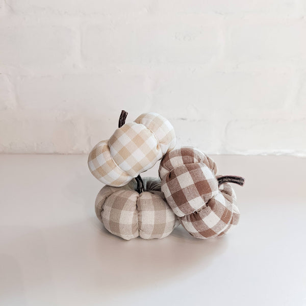Three mini pumpkin-shaped fabric pillows in checkered patterns of beige, white, and brown colors, arranged against a white brick wall background.