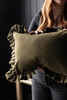 An olive green rectangular throw pillow with ruffled trim, held by a person in a dark room.