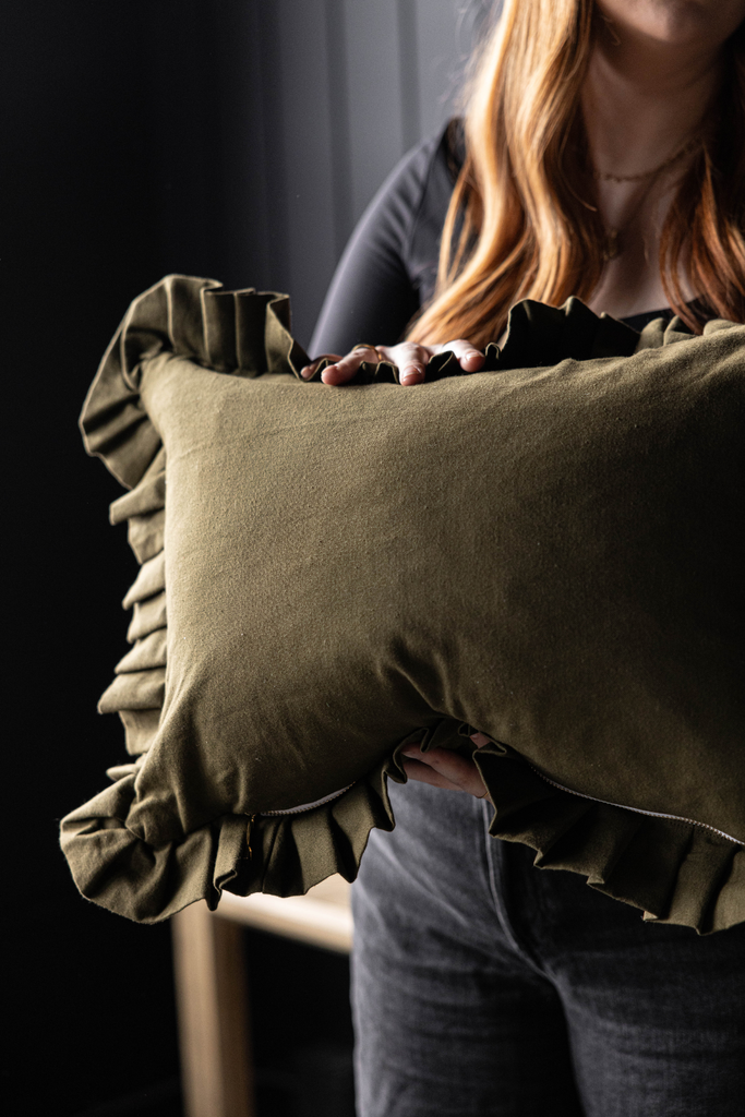 An olive green rectangular throw pillow with ruffled trim, held by a person in a dark room.