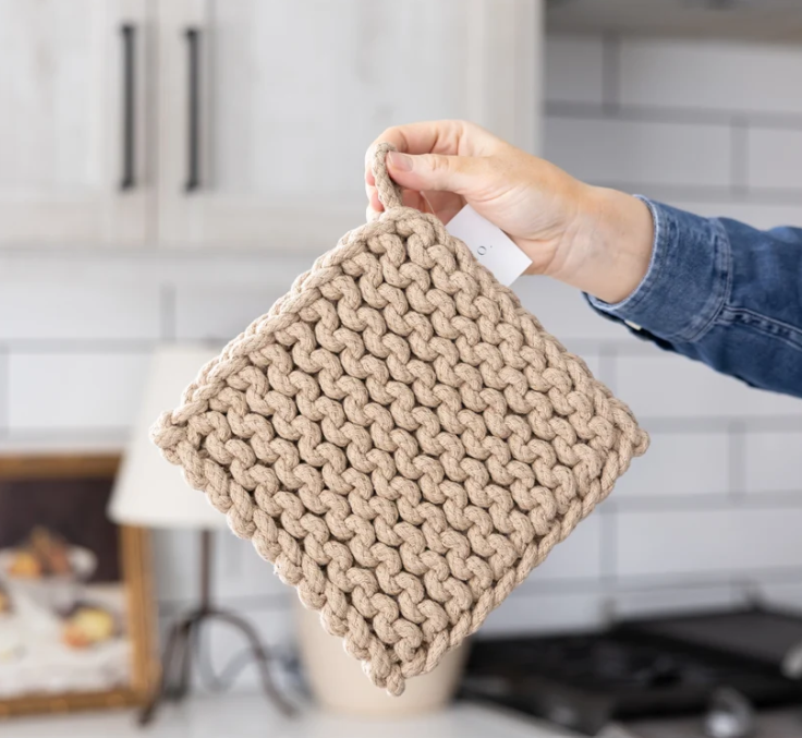 A hand holding a square, chunky-knit beige colored pot holder in a kitchen setting with white subway tiles and light wood cabinets in the background.