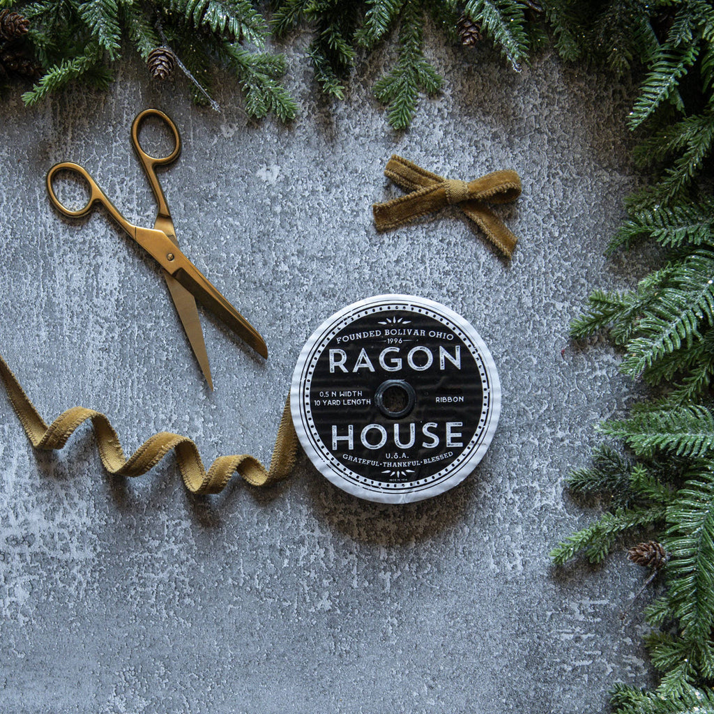 Ribbon spool with 'Ragon House' branding, gold scissors, and greenery on a textured surface