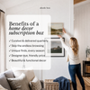 A woman in a white sweater and blue jeans hangs framed landscape art on a white wall in a bright living room. A large semi-transparent text box on the left reads 'Benefits of a home decor subscription box' with checkmarks listing curated quarterly delivery, no endless browsing, unique finds, designer eye, friendly price, and beautiful functional decor.