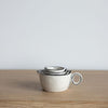 Three nested ceramic measuring cups with loop handles, featuring a warm off-white body and thin black rim detailing, displayed on a light wood surface against a neutral light gray background.