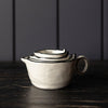 A set of nested ceramic measuring cups with a small handle and pouring spout, shown on a dark wooden surface against a dark paneled background.