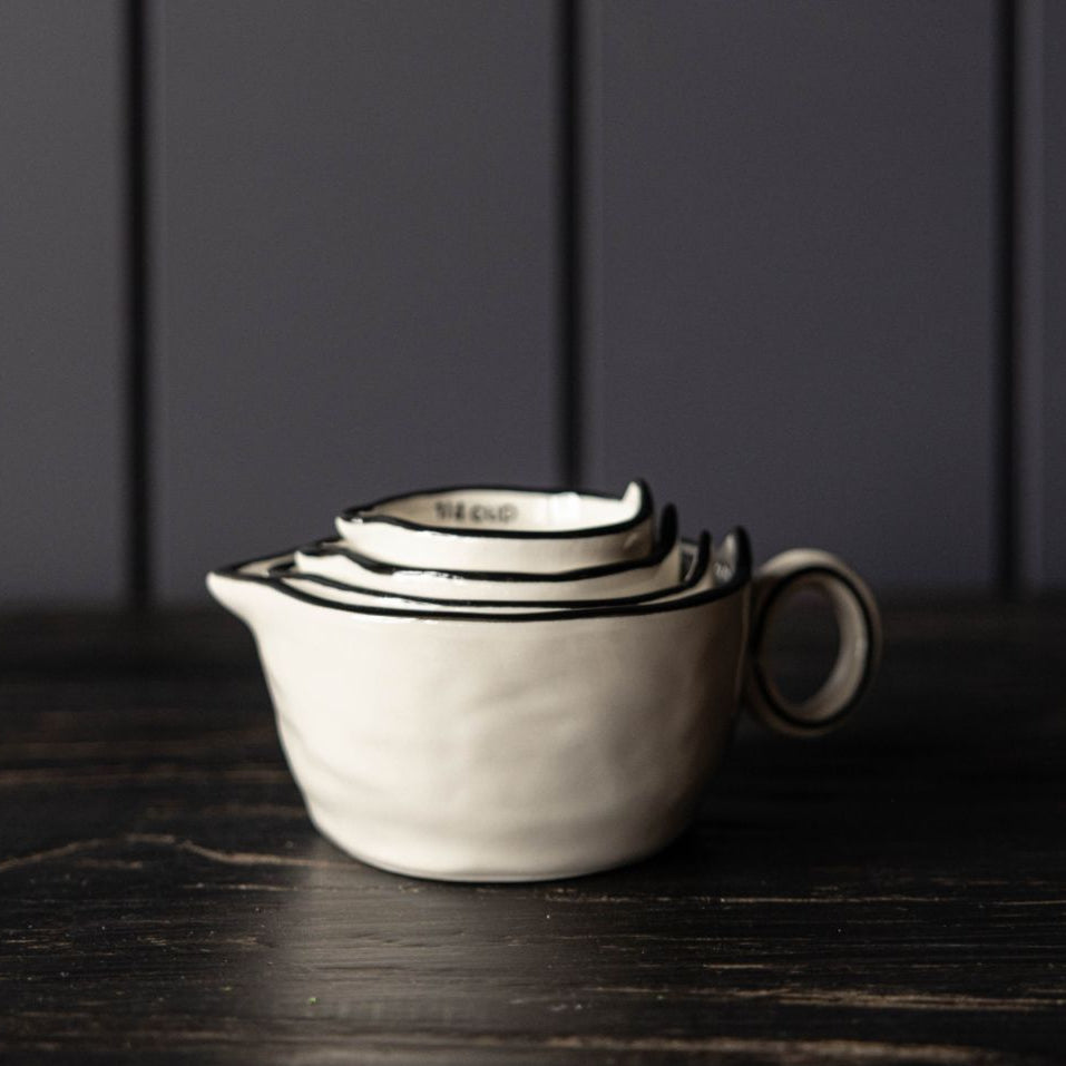 A set of nested ceramic measuring cups with a small handle and pouring spout, shown on a dark wooden surface against a dark paneled background.
