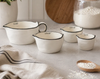 A set of four white ceramic measuring cups with black rim detailing and printed cup measurements, arranged on a light stone surface with a linen towel, a wooden spoon of flour, and a jar of flour in the background.