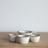 Four small ceramic measuring cups in different sizes, arranged on a light wood surface against a plain light gray background. The cups are off-white with black rim detailing and printed capacity markings including 1/4 cup, 1/2 cup, and 1/3 cup.