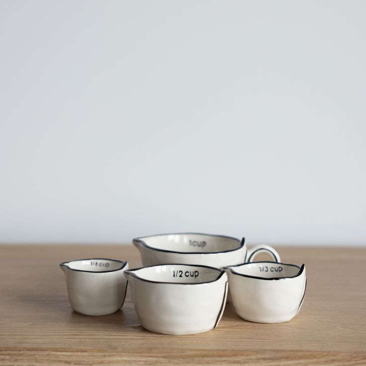 Four small ceramic measuring cups in different sizes, arranged on a light wood surface against a plain light gray background. The cups are off-white with black rim detailing and printed capacity markings including 1/4 cup, 1/2 cup, and 1/3 cup.