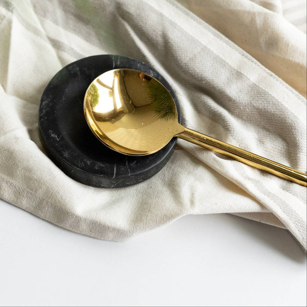 Gold spoon on a black stone spoon rest with a beige cloth and plant in the background