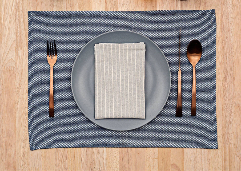 Dining table setting with gray plate, striped napkin, cutlery, and blue placemat on a wooden surface.