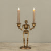 A skeleton-shaped double candle holder in an antique gold finish, holding two lit taper candles, displayed on a stone surface against a neutral gray background.