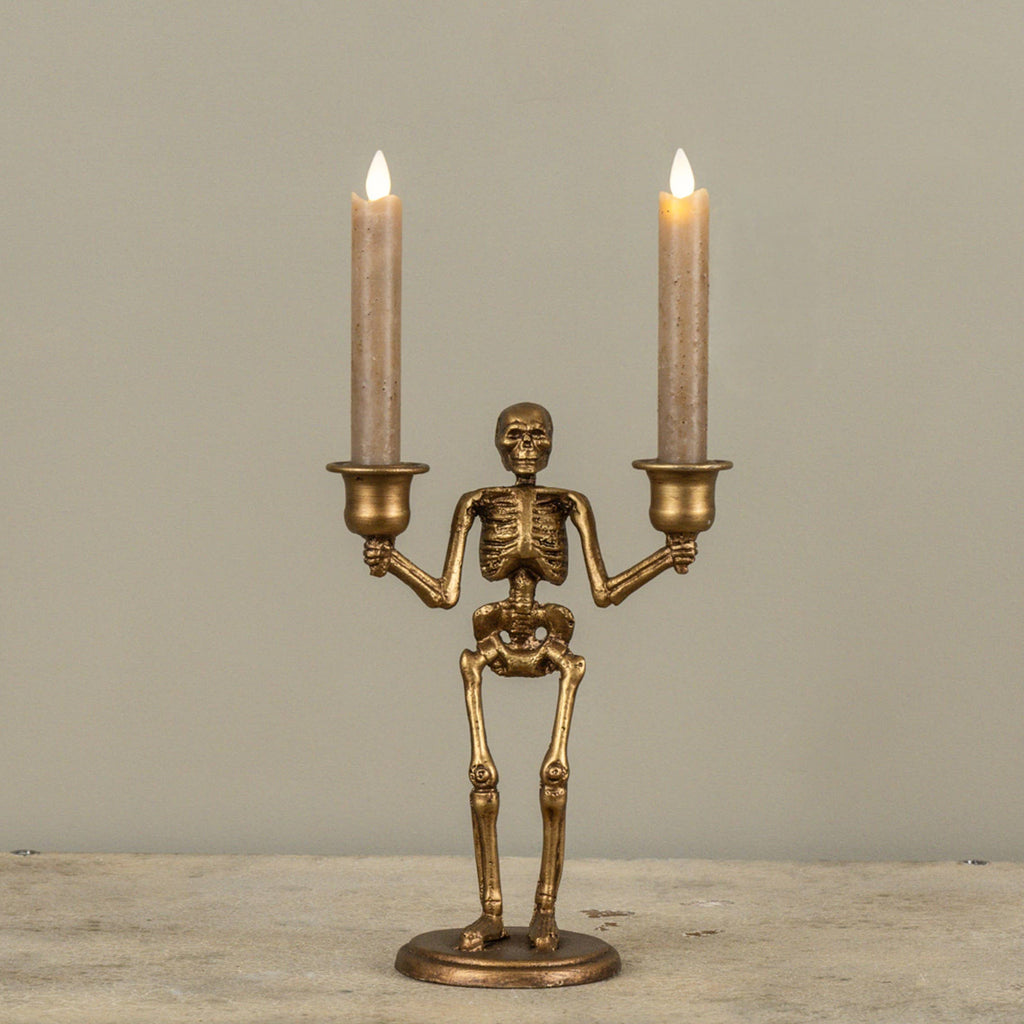 A skeleton-shaped double candle holder in an antique gold finish, holding two lit taper candles, displayed on a stone surface against a neutral gray background.