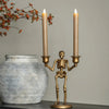 A gold skeleton candle holder with two lit taper candles sits on a dark wooden table next to a gray ceramic pot and fall foliage decorations.