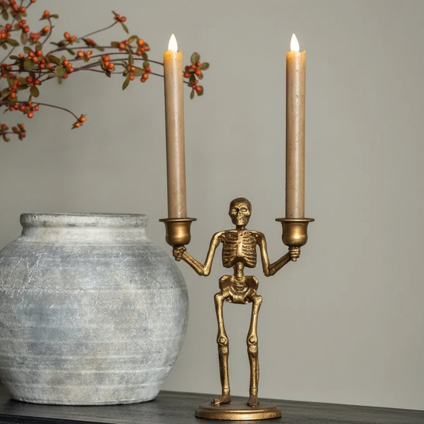 A gold skeleton candle holder with two lit taper candles sits on a dark wooden table next to a gray ceramic pot and fall foliage decorations.