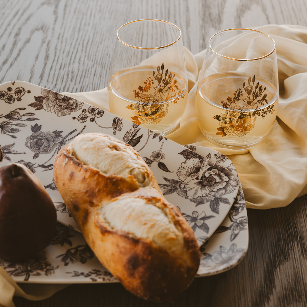 Two glasses of white wine with floral designs on a wooden table, accompanied by bread and pastries.