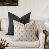 Two decorative throw pillows on a light beige sofa in a styled living room. The front pillow is cream with a small repeating dark gray botanical pattern, and the back pillow is dark gray with a subtle grid pattern.