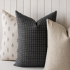Three decorative throw pillows arranged on a light upholstered bench against a white paneled wall. The center pillow is charcoal gray with a white windowpane grid pattern, flanked by two beige pillows with subtle woven textures and small black motifs.