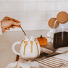 Person stirring a mug of hot chocolate with whipped cream and caramel drizzle, set against a cozy kitchen backdrop.