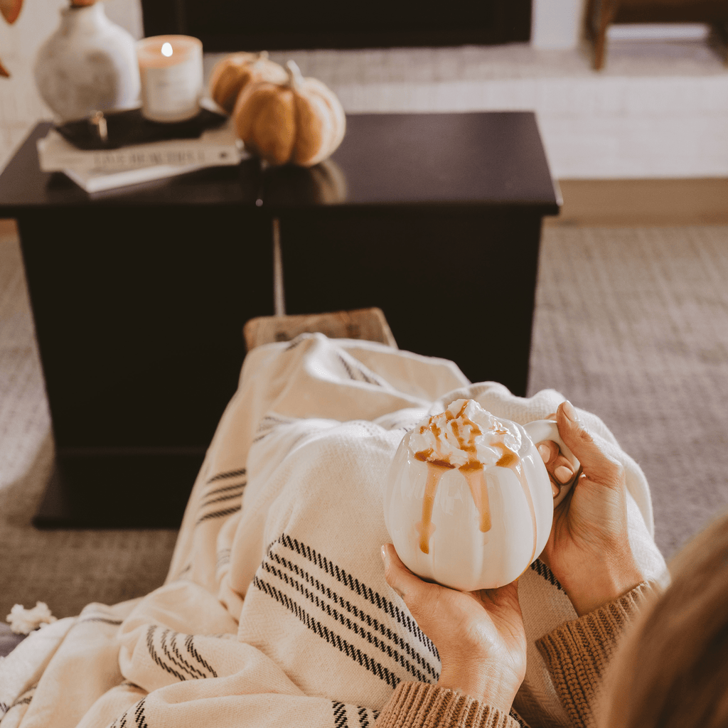 Person holding a small white pumpkin with brown drizzle, sitting on a couch in a cozy living room.