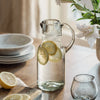 Clear glass pitcher with lemon slices on a wooden table with plates and flowers in the background