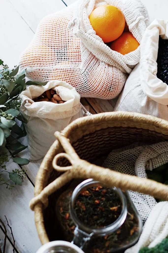 Mesh product bags with oranges and other items on a wooden surface, with a woven basket and teacup in the foreground.