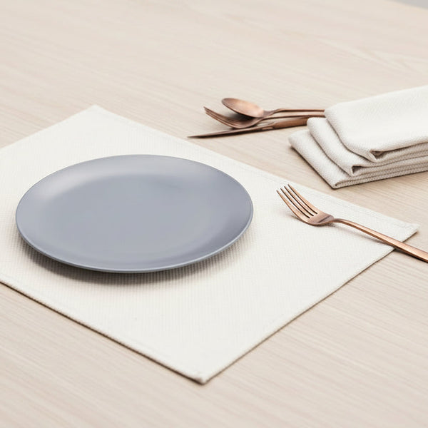 Gray plate on a cream cotton placemat with silverware and napkins on a light wooden surface