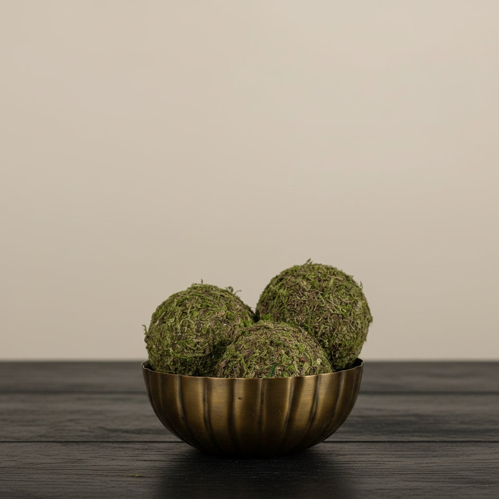Decorative arrangement with moss balls in a brass bowl, sunflowers, and a white vase on a wooden surface.
