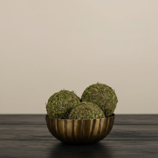 Decorative arrangement with moss balls in a brass bowl, sunflowers, and a white vase on a wooden surface.