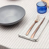 Dining table setting with a gray bowl, blue glass, wooden cutting board, and cutlery on a striped tablecloth.