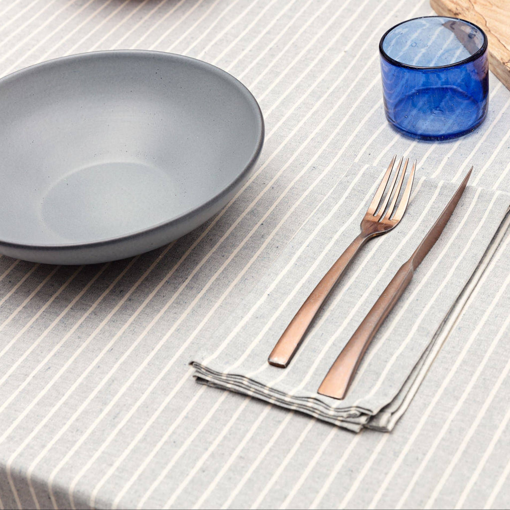 Dining table setting with a gray bowl, blue glass, wooden cutting board, and cutlery on a striped tablecloth.
