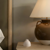 Brown ceramic lamp and small white bud vase on a wooden surface against a neutral wall.