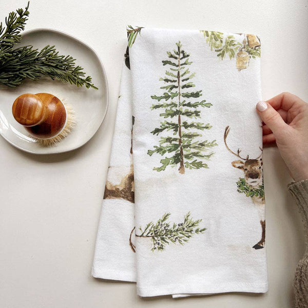 White towel with Christmas-themed design featuring trees and deer, held by a hand on a light surface.