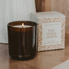 An amber glass jar candle with a lit flame sits on a wooden surface next to its decorative product box. The box is cream with a tan botanical pattern and reads 'FALL LEAVES' and 'Sweet Water Decor'.