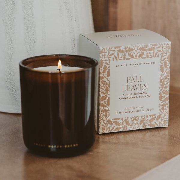 An amber glass jar candle with a lit flame sits on a wooden surface next to its decorative product box. The box is cream with a tan botanical pattern and reads 'FALL LEAVES' and 'Sweet Water Decor'.