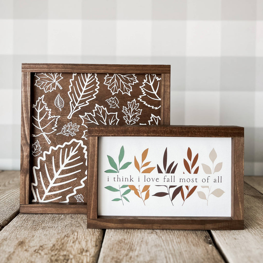 Two framed art signs with leaf patterns and the text 'I think I love fall most of all' displayed on a wooden surface.