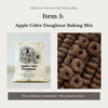 Promotional graphic for Modern Harvest Fall Abode Box's Apple Cider Doughnut Baking Mix. The image features a cream-colored drawstring bag with a black graphic of doughnuts and the product name, next to a close-up photo of a pile of baked apple cider doughnuts dusted with sugar.