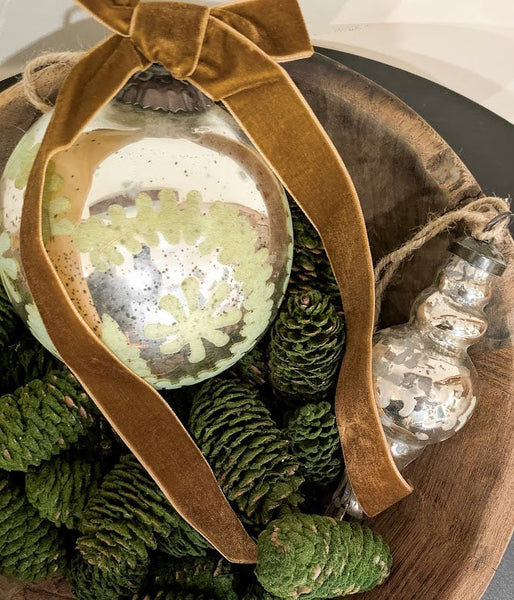 Decorative bowl with green ornaments, pinecones, and a gold ribbon on a neutral background