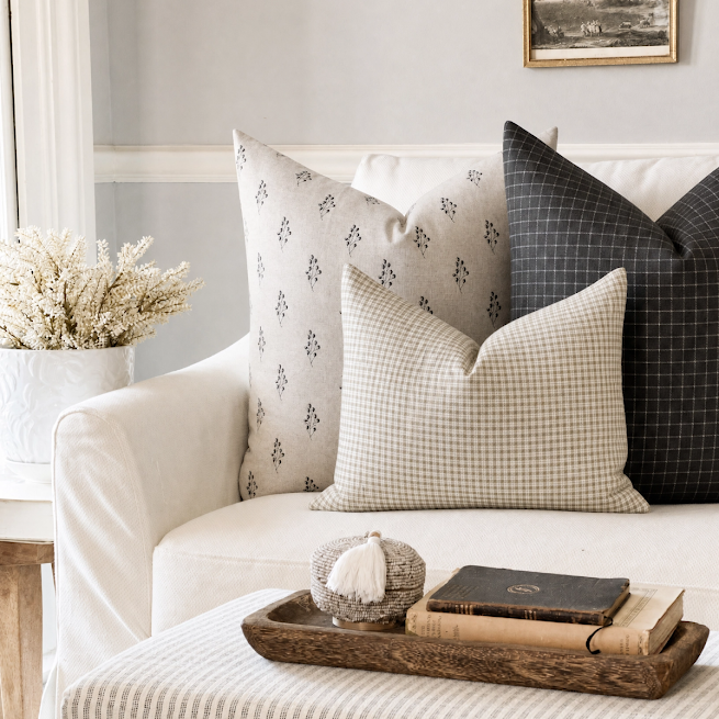 Three decorative throw pillows styled on a white sofa in a bright living room setting. The pillows include a cream pillow with a small dark geometric pattern, a cream and beige gingham check pillow, and a dark charcoal gray pillow with a subtle grid pattern.