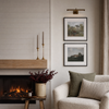 A cozy living room corner featuring a cream-colored upholstered armchair with green and striped throw pillows, a rustic wooden side table with a vase of burgundy foliage, and a fireplace with a light wood mantel. Two framed landscape prints hang on the wall above the chair.