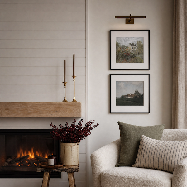 A cozy living room corner featuring a cream-colored upholstered armchair with green and striped throw pillows, a rustic wooden side table with a vase of burgundy foliage, and a fireplace with a light wood mantel. Two framed landscape prints hang on the wall above the chair.