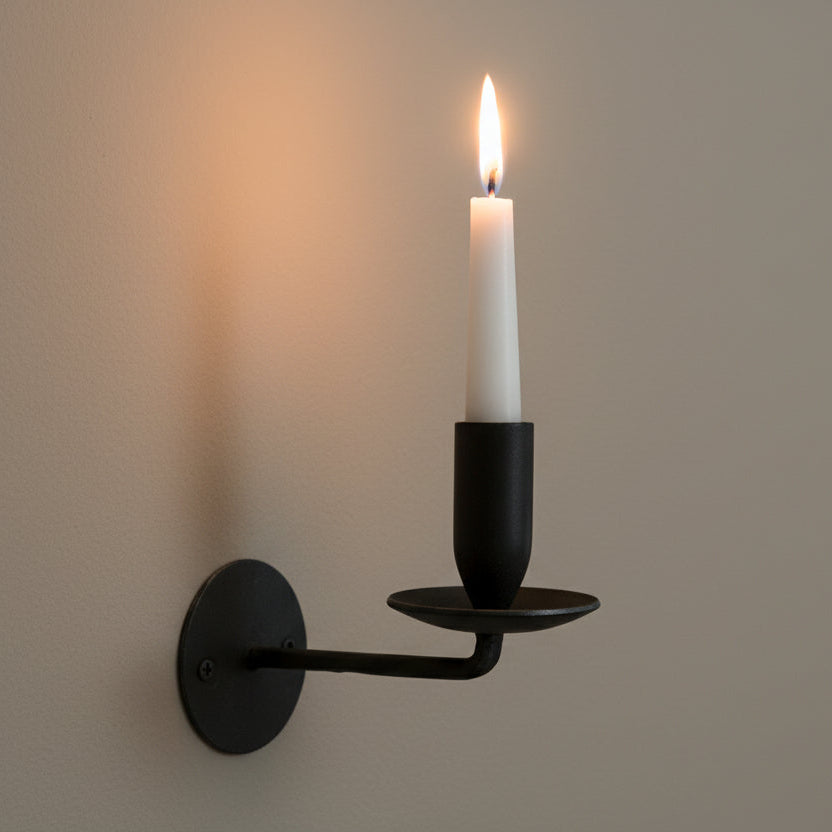 Black wall-mounted candle holder with a lit candle on a plain wall.