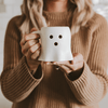 Person holding a ghost shaped coffee mug with black dots, wearing a brown sweater.