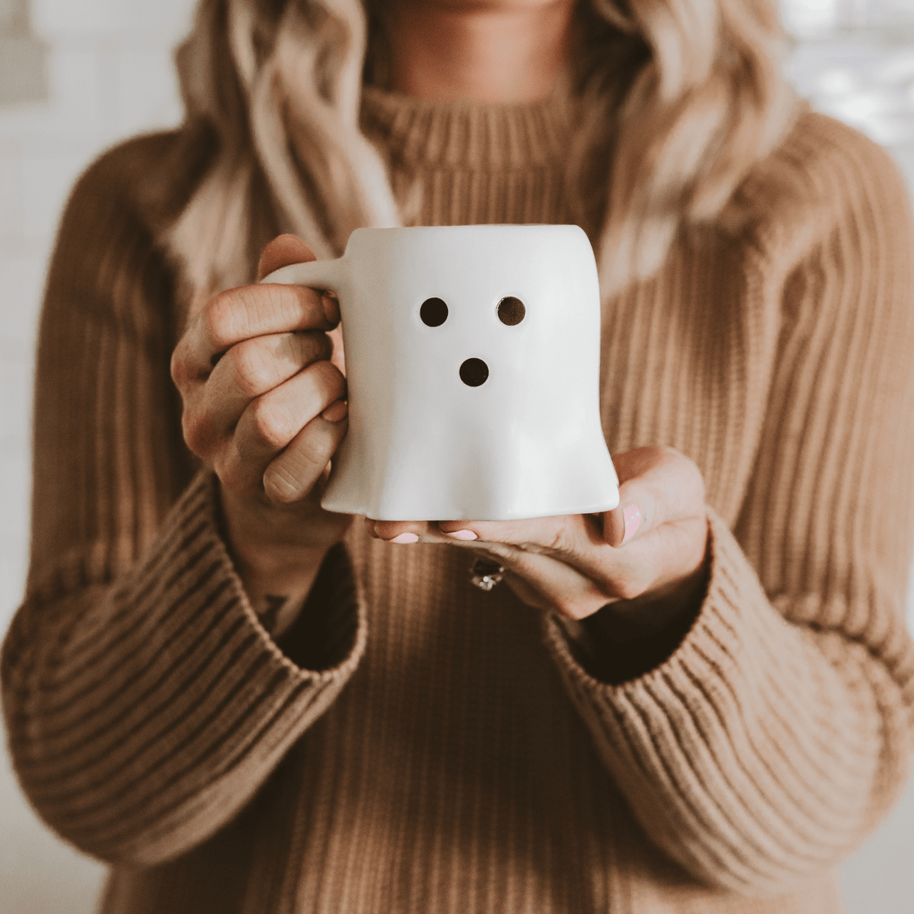 Person holding a ghost shaped coffee mug with black dots, wearing a brown sweater.