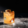 An orange-hued cocktail in a clear glass tumbler with two small white ghost decals, garnished with a sugar cube and orange peel, set against a dark smoky background.