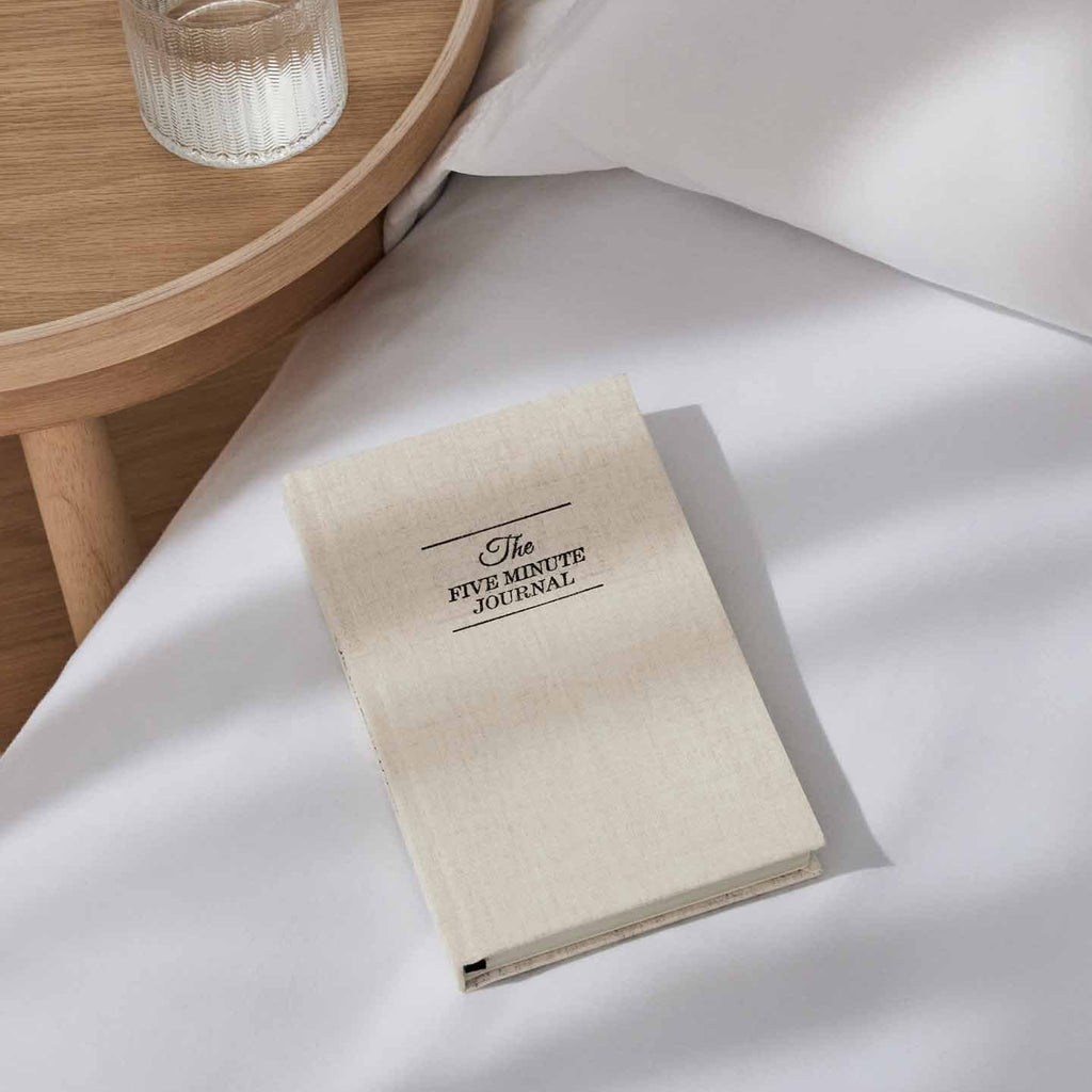Beige notebook titled 'The Five Minute Journal' on a white surface with a wooden side table in the background.