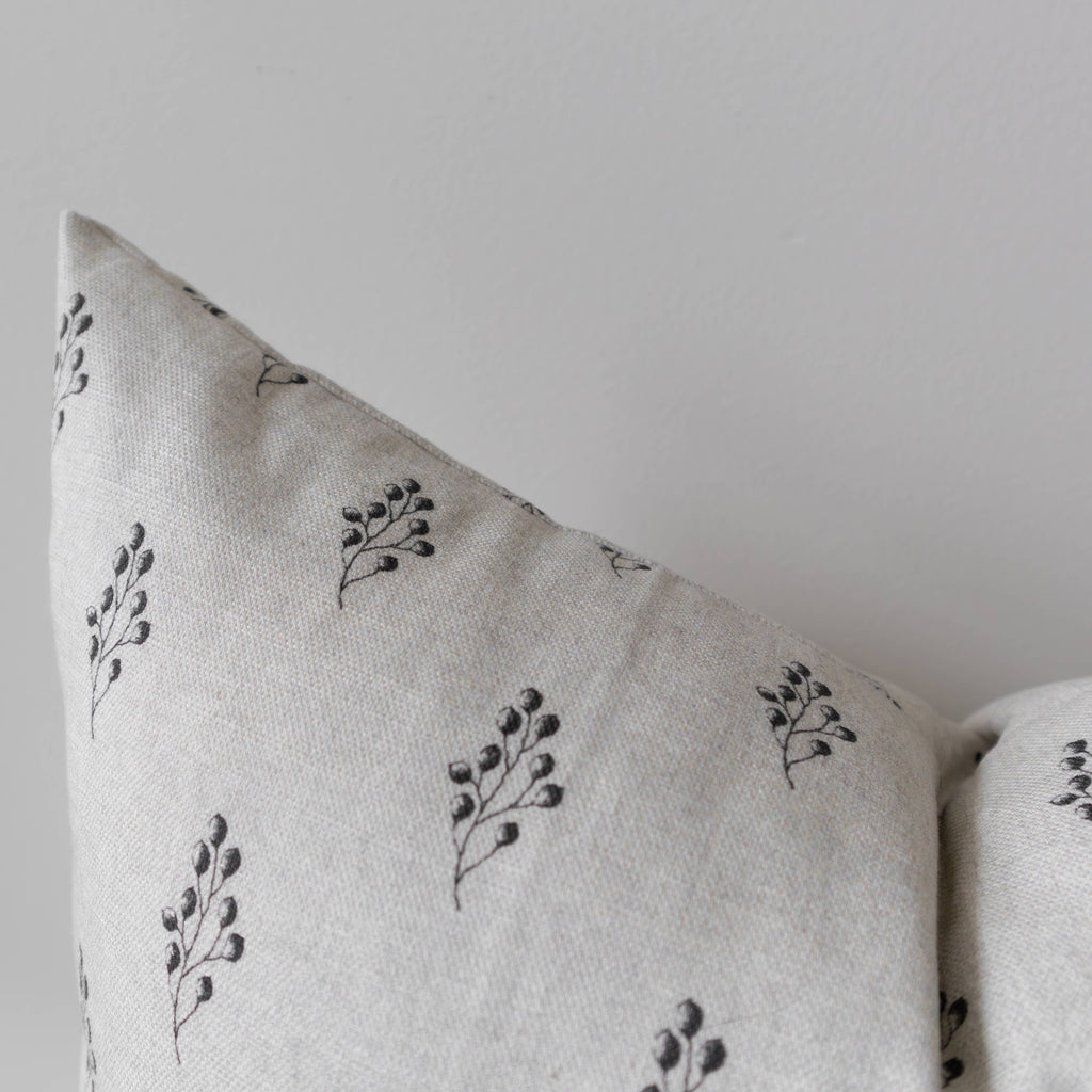 A close-up of a light beige linen throw pillow with a delicate black botanical leaf pattern, shown against a neutral light gray background.