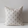A square throw pillow in a light beige linen fabric with a repeating black floral block print pattern, resting on a white surface against a neutral gray background.