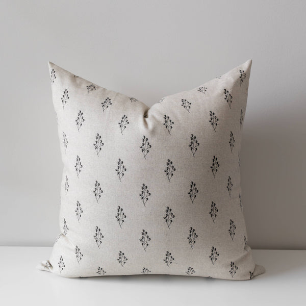 A square throw pillow in a light beige linen fabric with a repeating black floral block print pattern, resting on a white surface against a neutral gray background.